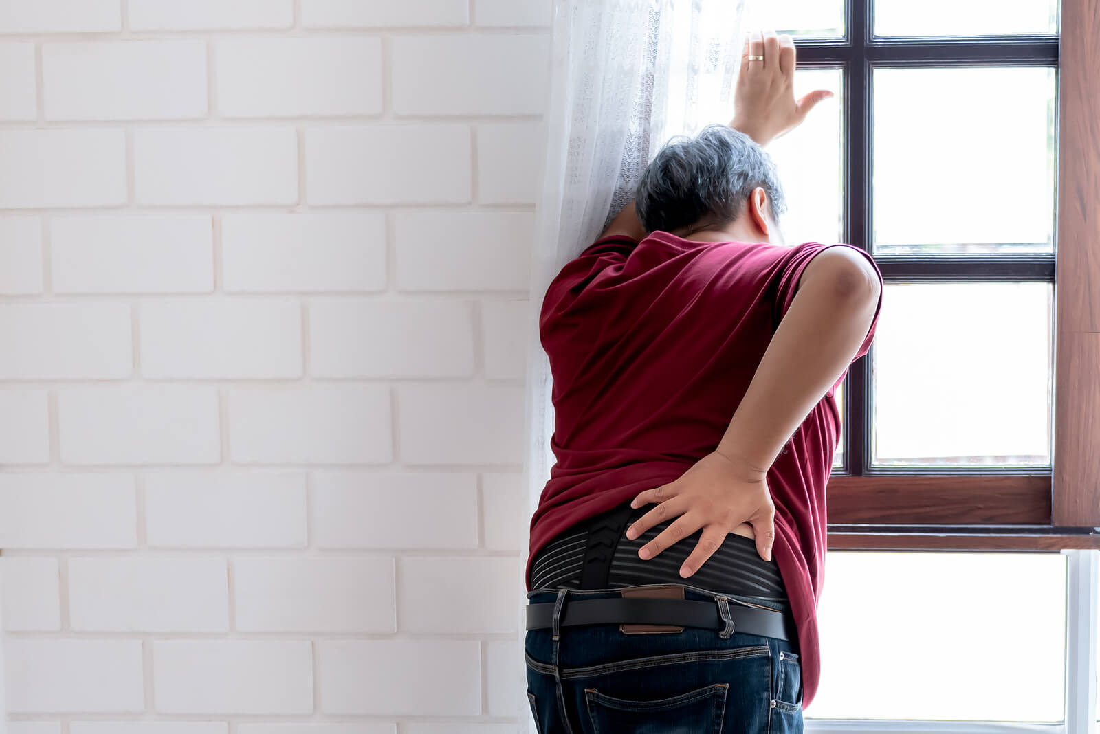 A person leans on a window they are looking out of. They are holding their lower back. Learn more about chronic illness counseling by contacting a chronic illness therapist in New Bern, NC. We offer pain reprocessing therapy in New Bern, NC and other services.