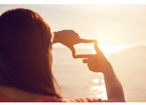 Woman looking at the sunset over the ocean with her hands framing the scene like a photo. Online therapy in North Carolina can help you process the past with the best online therapist. Improve your mental wellness with life changes counseling, anxiety treatment, trauma therapy and more!
