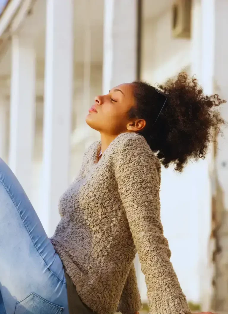Relaxed young woman with natural curly hair enjoying sunlight, eyes closed, wearing cozy sweater and jeans outdoors.