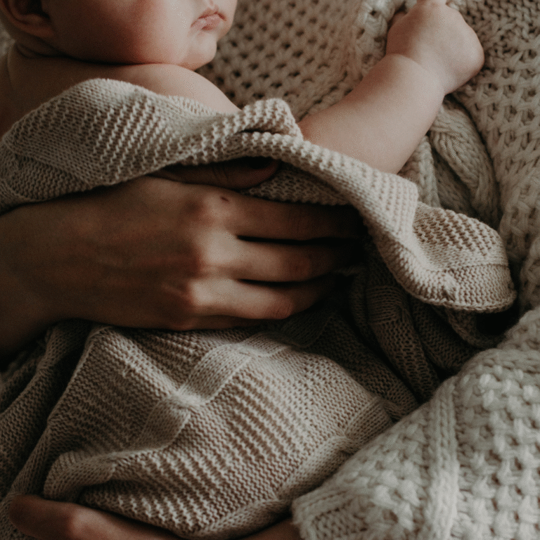 Newborn wrapped in a soft knit blanket, held closely - representing the tenderness and vulnerability of early postpartum moments.