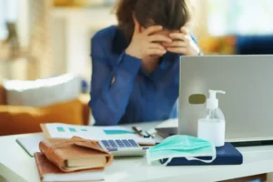 Stressed woman at desk with laptop, face mask, and sanitizer.