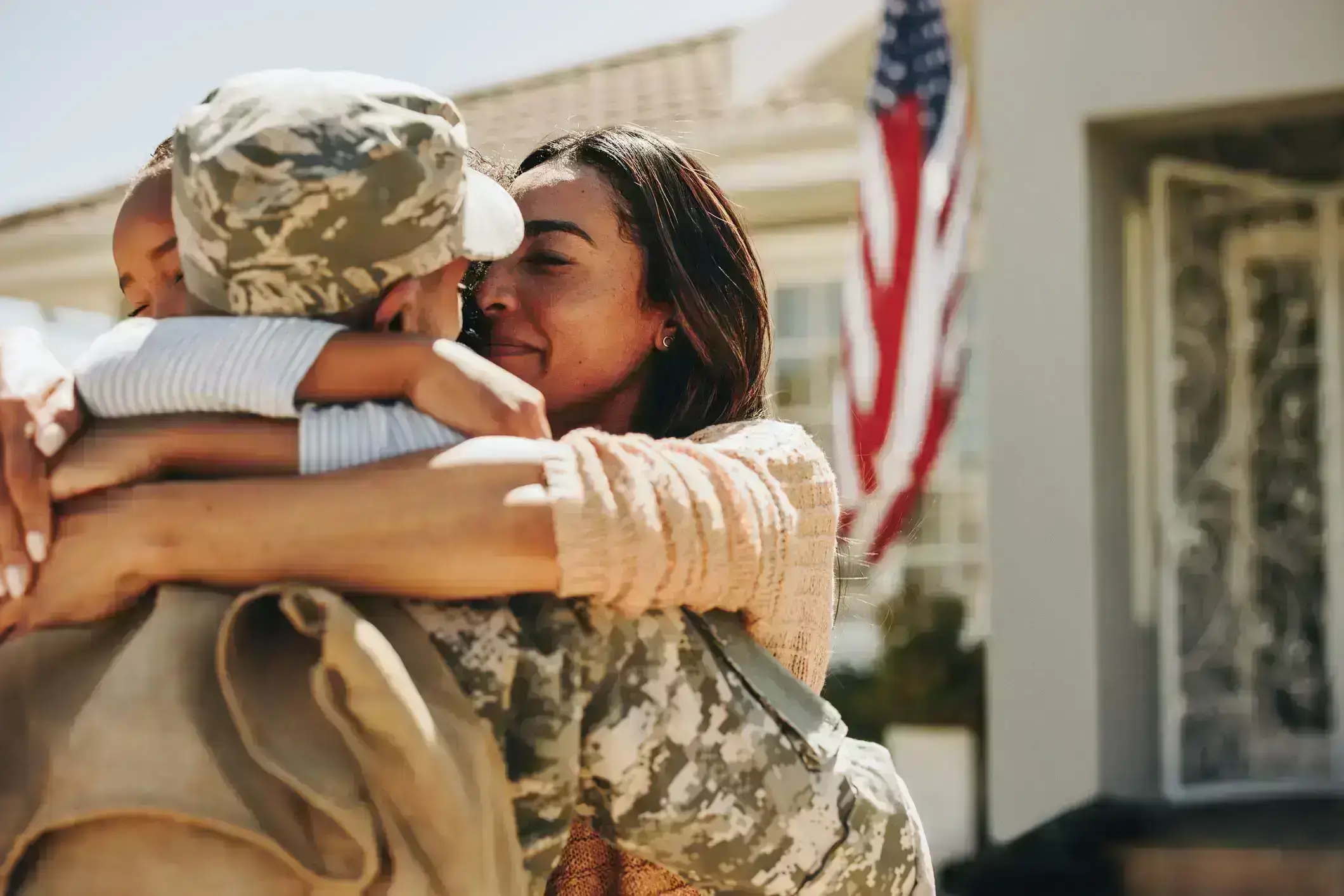 Soldier reunites with family in heartfelt hug outside home, with American flag showing patriotism and pride.