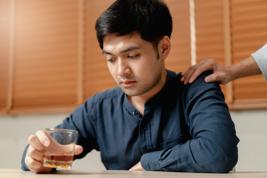 Young man sitting at table with alcohol, friendly hand on his shoulder in support