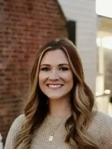 Kaitlyn Moore, LCSW therapist at Renewed Wellness Counseling, smiling outdoors in front of a brick and white building, wearing a beige sweater and gold pendant necklace.