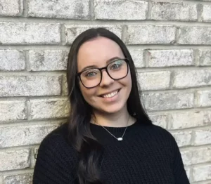 Savannah Reed, LCSW therapist at Renewed Wellness Counseling, smiling in front of a light brick wall, wearing glasses and a black textured sweater with a simple pendant necklace.
