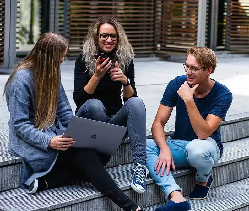 Three young adults sitting on steps outside, talking and smiling with a laptop and phone — illustrating peer support and managing stress in a social setting.