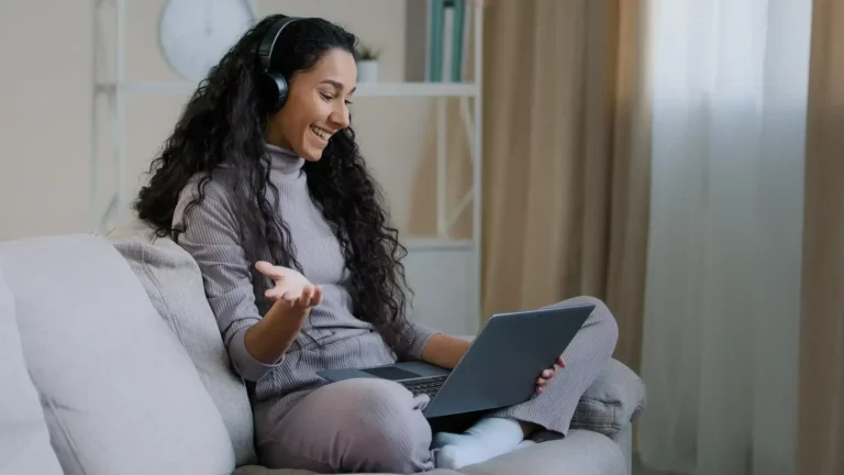 Young hispanic arab attractive woman freelancer with curly hairstyle wear headset sit in lotus pose on cozy sofa hold laptop talk distance wireless connection undergoing psychotherapy session remotely