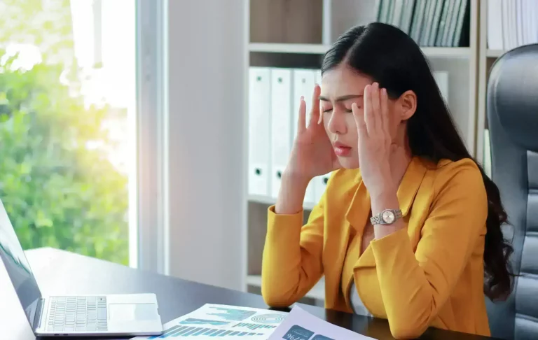 Stressed businesswoman sitting at desk with eyes closed and hands on temples, showing signs of workplace anxiety.