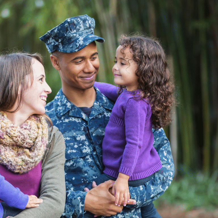 Military father in uniform smiling and holding his young daughter, with his wife standing beside them, all sharing a warm family moment outdoors.