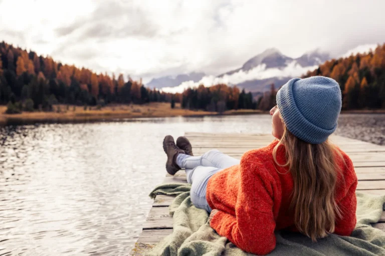 Woman in warm clothes relaxing on a dock by a lake, surrounded by autumn trees and mountains in the distance.