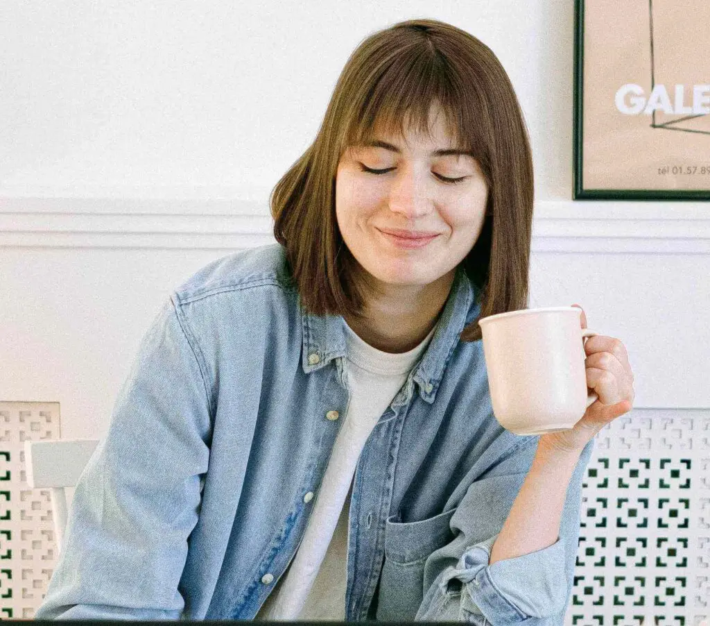Smiling woman in a denim shirt holding a mug, sitting indoors and appearing content.