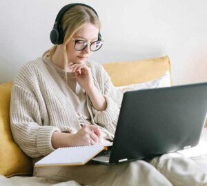 Woman with headphones taking notes while using a laptop, sitting on a couch.