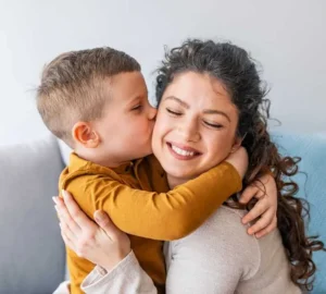 Young boy kisses his smiling mother on the cheek as they share a warm hug on the couch.