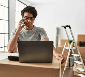 Man talks on phone while working on laptop during a home move, surrounded by boxes and a step ladder.