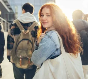 Red-haired woman smiling back while walking in sunlight, wearing a denim jacket and carrying a canvas tote bag.