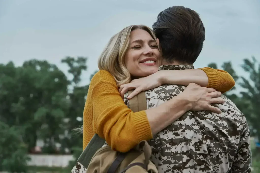 Smiling woman hugging a soldier in camouflage uniform outdoors.