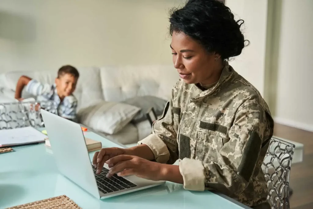 Woman in military uniform working on a laptop at home with child in background.
