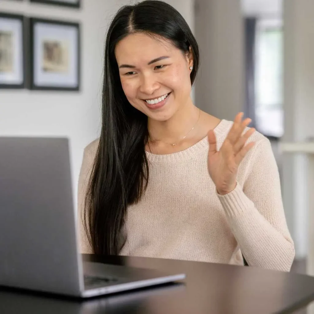 Woman smiling and waving during online therapy session via laptop at home.