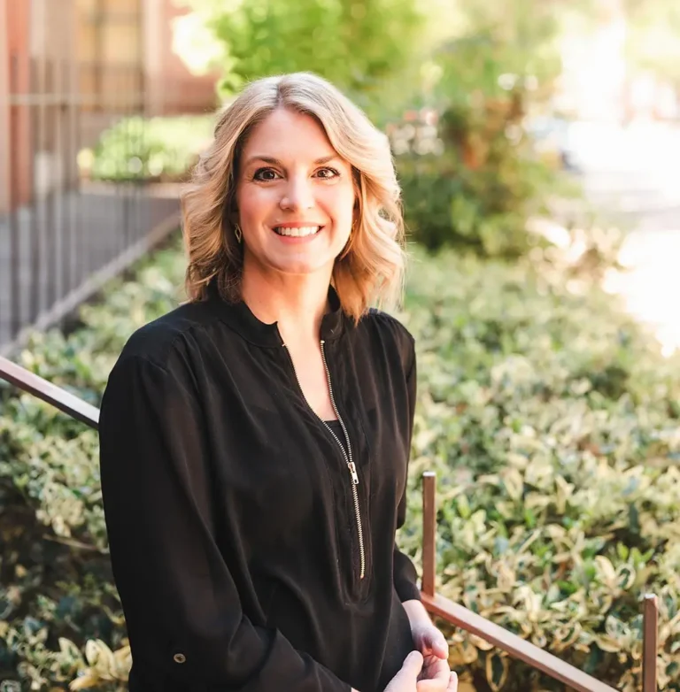 Marcy Arnold, LCSW, LISW-CP, LCAS, smiling outdoors in professional attire, standing in front of greenery and a sunlit walkway