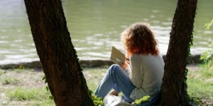 Woman with curly hair reading a book by a peaceful lake under trees, practicing self-care and relaxation during summer — Renewed Wellness Counseling, New Bern NC