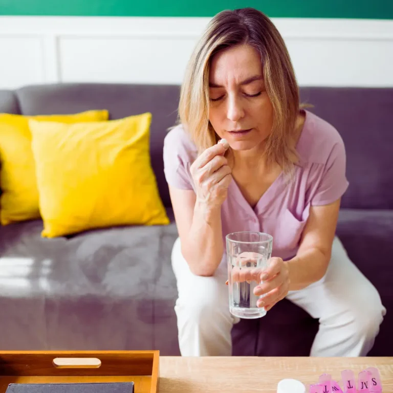Woman sitting on a couch taking medication with a glass of water, managing chronic illness and self-care at home — Renewed Wellness Counseling, New Bern NC