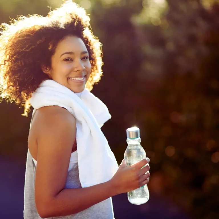 Smiling woman with curly hair holding a water bottle and towel, practicing self-care and hydration during summer exercise — Renewed Wellness Counseling, New Bern NC