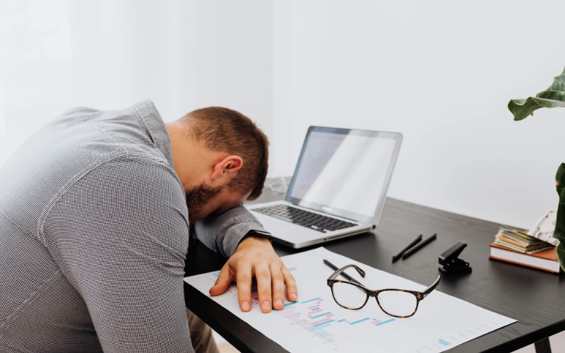 Man asleep at desk with head down near laptop, showing fatigue from poor sleep and mental burnout