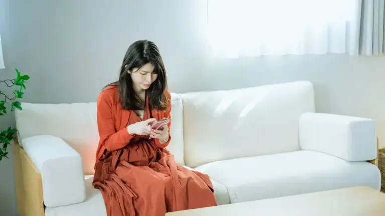 Woman sitting alone on a couch looking at her phone, appearing anxious and withdrawn, reflecting internal stress and overthinking linked to trauma