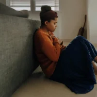 Person sitting on the floor leaning against a couch with hands on their chest, illustrating a quiet moment of overwhelm and the need for emotional support.